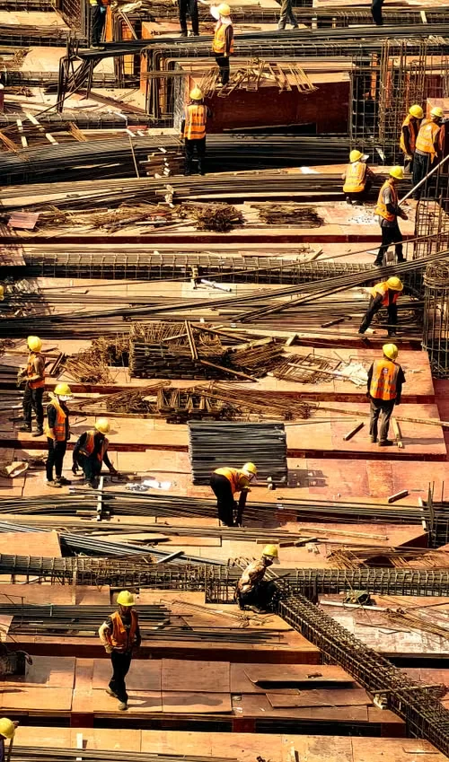 Vue plongeante d’ouvriers en casques et gilets orange parmi les armatures et coffrages d’un chantier.